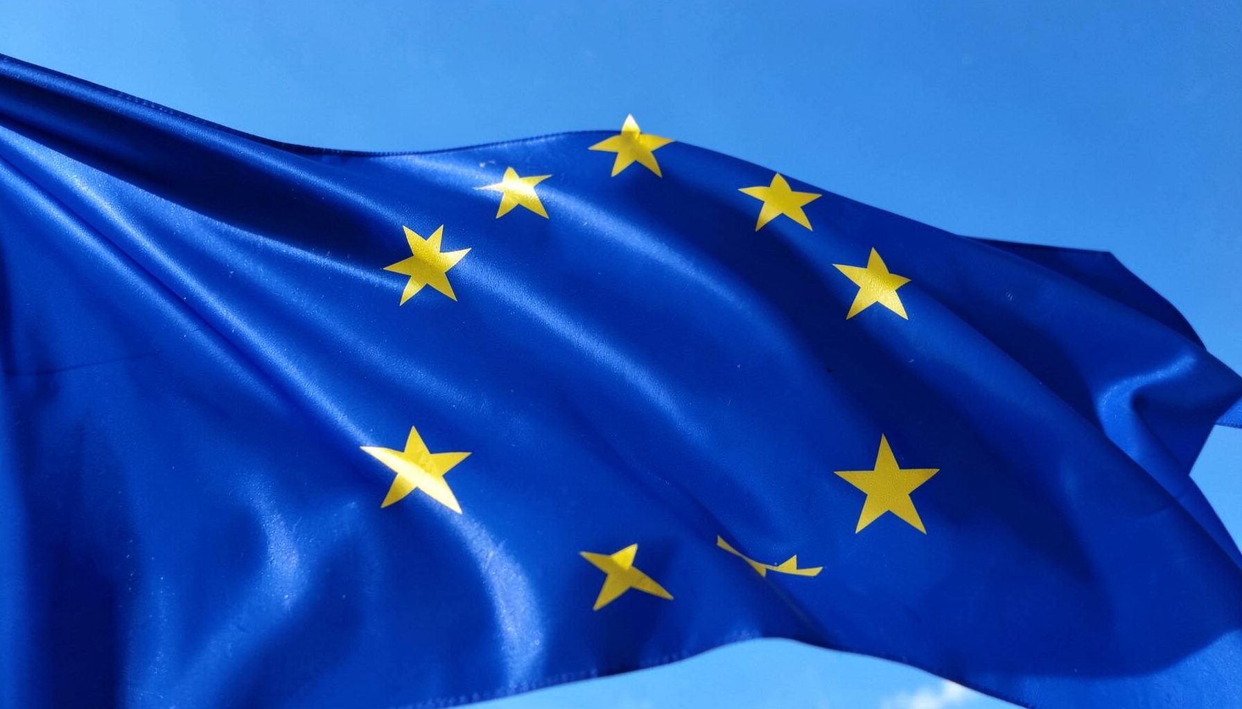 A close-up of the European flag rippling gently against a clear blue sky, the twelve gold stars catching bright sunlight