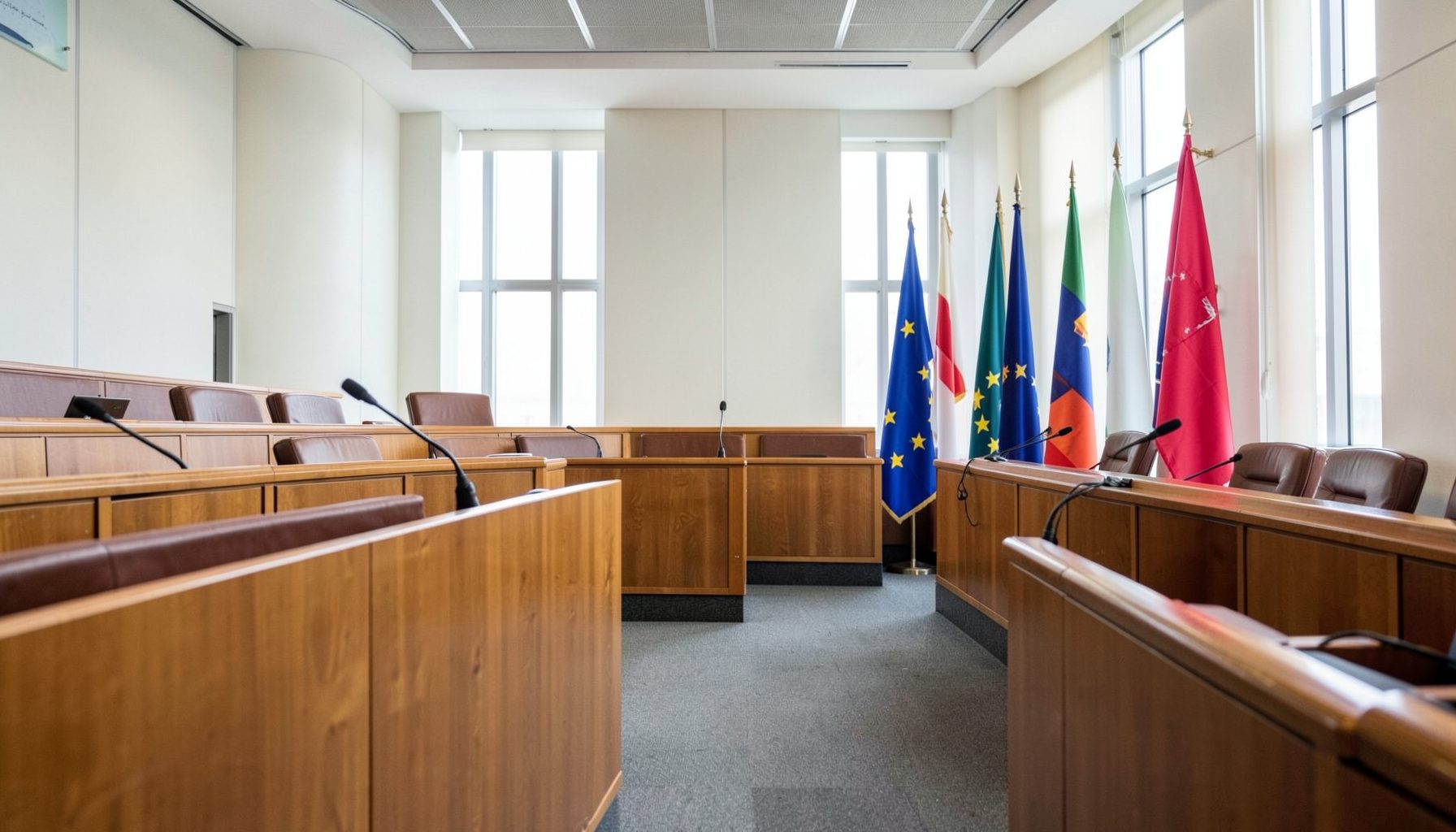 A modern courtroom interior with wooden benches, microphones, and the European flag standing alongside national flags, lit by natural daylight